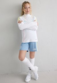 Witte sportieve top met lange mouwen en roze accenten, gecombineerd met lichtblauwe shorts met een logo, witte kniehoge sokken en witte sportschoenen.