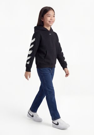 Jaunas žmogus vaikšto vilkėdamas juodą gobtuvą su baltomis juostelėmis ant rankovių, mėlynais džinsais ir baltais Nike sportbačiais ant balto fono.