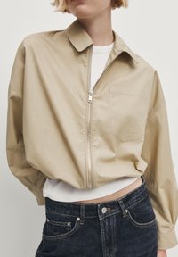 Beige lightweight jacket with a zipper, a collar, and a chest pocket. Paired with a white ribbed shirt and dark blue denim jeans.