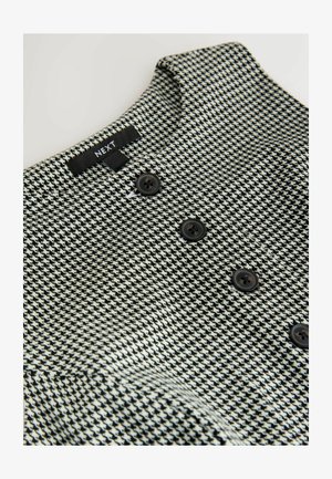 Tissu à motif pied-de-poule noir et blanc avec un col montant, comportant trois boutons foncés et une étiquette portant la mention "NEXT."