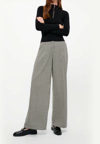 Kvinna som står med armarna i kors, iförd en svart zip-up topp, svartvita houndstooth byxor med vida ben och svarta loafers med guld detaljer.