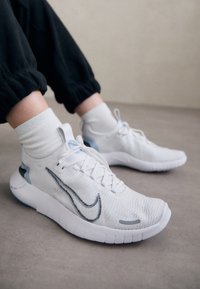 Zapatillas deportivas blancas con parte superior de tejido transpirable, logo Swoosh negro cosido, detalles azules en el talón y diseño de suela acolchada.