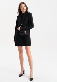 Czarny zestaw marynarki i spódnicy, wykonany z gładkiego materiału. Bluzka w kropki pod spodem. Czarna torebka z imitacji krokodyla z złotymi akcentami. Czarne szpilki.
