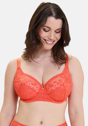 Femme portant un soutien-gorge corail en dentelle armuré, regardant vers le bas et souriant, avec des cheveux bruns mi-longs sur un fond uni.