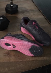 Ζευγάρι μαύρα και ροζ αθλητικά παπούτσια Adidas τοποθετημένα σε ξύλινο πάτωμα δίπλα σε δύο μαύρα εξαγωνικά αλτήρες 25 λιβρών.