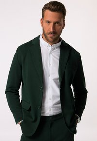 Blazer verde scuro realizzato in tessuto morbido, con colletto a punta, due bottoni e una tasca sul petto, abbinato a una camicia bianca.