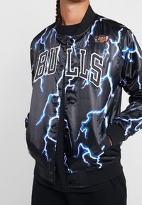 Musta satiininen bomber-takki, jossa siniset salamaniskut, "BULLS"-logo ja NBA-finaalien merkki. Sisältää resorit hihansuissa ja taskut.