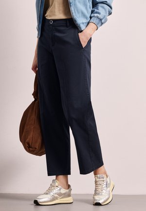 Person iført marineblå bukser med vide ben, sølvfarvede sneakers, lyseblå jakke, holder en brun taske med den ene hånd i lommen mod en ensfarvet baggrund.