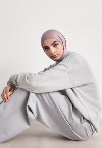 Grauer Strickpullover und hellgraue weite Hose, getragen von einer Person, die mit den Armen die Beine umwickelt sitzt und ein rosa eng anliegendes Kopftuch trägt.