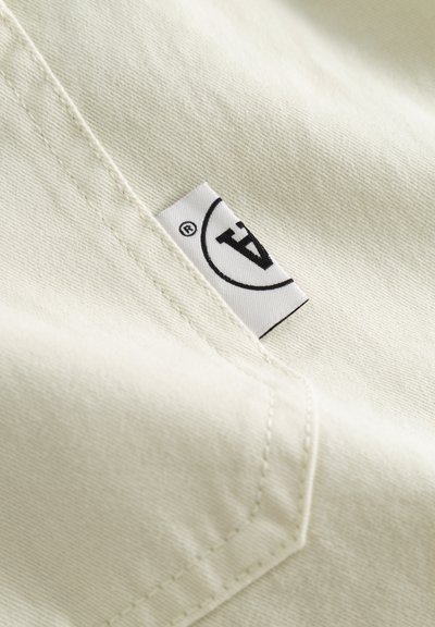 Gros plan sur un tissu crème avec des coutures visibles et une petite étiquette blanche portant un logo circulaire noir et la lettre "A".