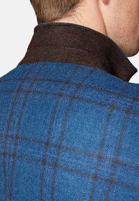 Niebieska plażowa kurtka wełniana w kratę z ciemnobrązowym filcowym kołnierzem; charakteryzuje się strukturą tkaniny i kontrastującym przeszyciem wzdłuż krawędzi kołnierza.