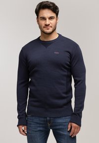 Granatowy sweter dzianinowy z okrągłym dekoltem, ściągaczami na mankietach i u dołu. Posiada mały akcent w postaci logo na klatce piersiowej oraz teksturowany wzór.