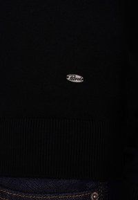 Maglione nero a maglia con orlo a costine e un logo ovale argentato sul petto. Abbinato a jeans scuri visibili in fondo.