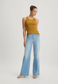 Canottiera marrone a coste con un logo, abbinata a jeans a gamba larga di un blu chiaro, con una leggera scoloritura e una vestibilità rilassata, indossata con sandali con cinturini.
