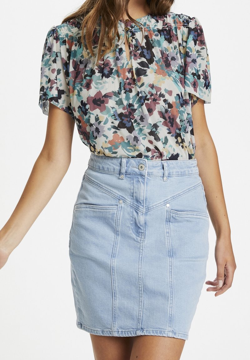 Femme portant un chemisier fleuri à manches courtes, rentré dans une jupe en jean bleu clair avec des poches avant et des coutures visibles.