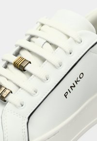 Λευκό δερμάτινο αθλητικό παπούτσι με υφή στην επιφάνεια, λευκά κορδόνια και χρυσαφένιες λεπτομέρειες στα κορδόνια. Το όνομα της μάρκας "PINKO" σε μαύρο χρώμα.