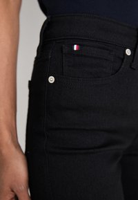 Jeans de mezclilla negra de cintura alta, con herrajes metálicos, cinco bolsillos y un pequeño acento de logo rojo, blanco y azul en el bolsillo trasero.