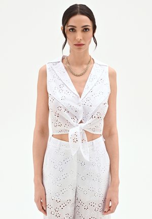 Femme aux cheveux foncés attachés, portant un haut sans manches blanc en broderie anglaise avec un nœud sur le devant et un pantalon assorti, accessoirisée avec un collier chaîne en or.