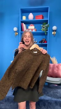 Woman with long blonde hair smiling, holding a brown sweater on a hanger in a blue room with shelves, books, plants, and decorative items.