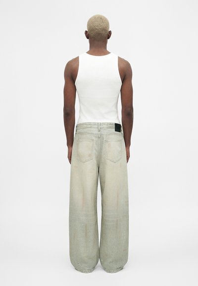 Άτομο με κοντά ξανθά μαλλιά που φοράει ένα λευκό τοπ χωρίς μανίκια και πλατιά ξεβαμμένα ανοιχτόγκομα τζιν, φαίνεται από πίσω.