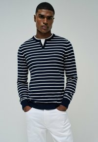 Pullover a righe navy e bianche in maglia con scollo henley, dotato di bottoni e polsini a costine, abbinato a pantaloni bianchi.