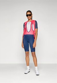 Equipamento de ciclismo com um colete de malha rosa e calções almofadados azuis. Ambos são justos e têm logótipos, combinados com óculos de sol e sapatos brancos.