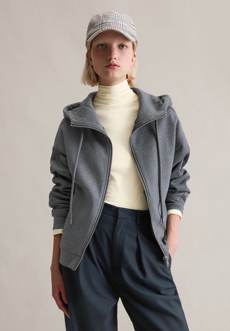 Grijze hoodie met rits en capuchon, voorzien van geribbelde manchetten en trekkoorden, gedragen over een lichtgele coltrui. Model gecombineerd met marineblauwe broek.