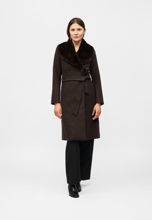 Une femme porte un manteau marron foncé ceinturé à col douillet, un pantalon noir et des bottines, sur un fond clair uni.