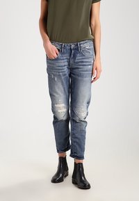 Personne portant un jean bleu délavé et usé avec des manches retroussées, une chemise oliver à manches courtes, et des bottines noires, se tenant debout contre un fond uni.
