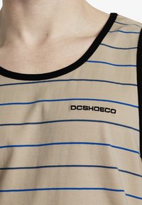 Camiseta sin mangas beige con rayas horizontales negras y azules, con un escote acanalado negro y el logo "DCSHOECO" bordado en el pecho.