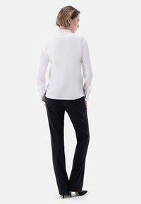 Blusa bianca a maniche lunghe con colletto, abbinata a pantaloni neri e scarpe con tacco alto, vista da dietro su uno sfondo chiaro.