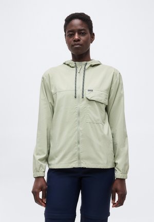 Person wearing a light green hooded zip jacket with front pocket and navy pants, standing against a plain white background.