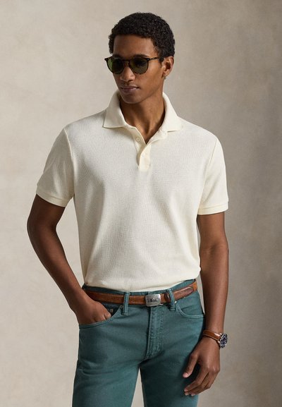 Cream-colored, textured polo shirt with a collared neckline, paired with teal jeans and a brown leather belt. Circular, dark sunglasses accessorize.