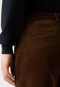 Brune corduroy bukser med et struktureret ribbet mønster og detalje med en baglomme, parret med en mørk trøjesleeve.