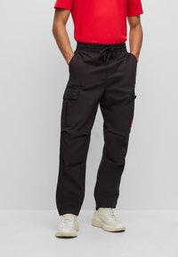 Black cargo pants with an elastic waistband and drawstring. Features multiple side pockets and a relaxed fit. Worn with a red t-shirt and light sneakers.