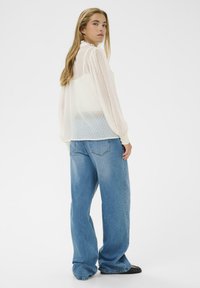 Hvid bluse med prikker og puffede ærmer, parret med lyseblå løstsiddende jeans, sat mod en ensfarvet hvid baggrund.