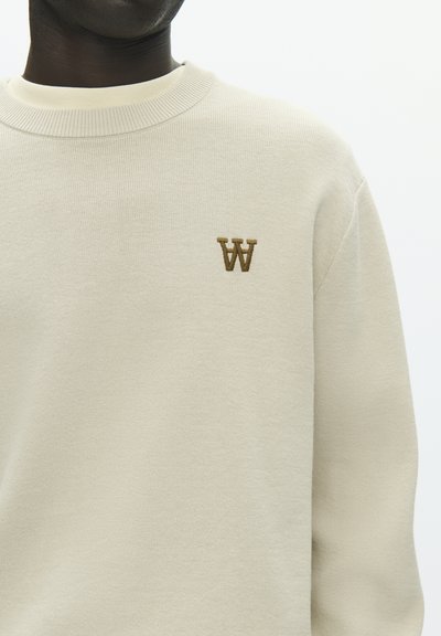 Sweat-shirt couleur crème en matière douce, avec un col côtelé et un petit "W" brun brodé sur la poitrine.
