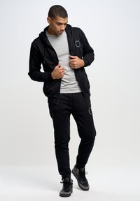 Schwarze Zip-Hoodie mit Taschen über einem grauen T-Shirt, kombiniert mit schwarzen Jogginghosen mit einem Logo. Getragen mit grauen und schwarzen Turnschuhen.