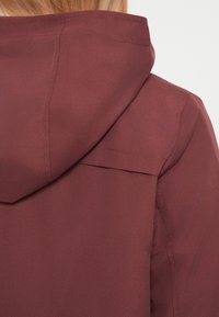 Κοντινό πλάνο ενός ατόμου που φοράει σκούρο κόκκινο μπουφάν με κουκούλα, δείχνοντας την πλάτη και τις λεπτομέρειες της κουκούλας με ορατή ραφή.