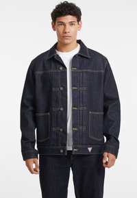 Dark blue denim jacket with a buttoned front, two chest pockets, and contrasting yellow stitching. Features a classic collar and relaxed fit.