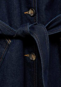 Vêtement en denim foncé avec une ceinture nouée, des boutons marron et des coutures orange contrastantes. Le tissu a une texture lisse et un design classique.