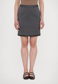 Donna che indossa una gonna grigia a righe con bordi neri, una maglia marrone e scarpe flat a doppia cinghia marrone scuro, in piedi contro uno sfondo bianco.