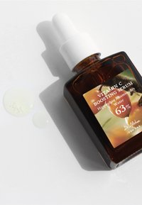 Botella de vidrio ámbar con un gotero blanco, etiquetada "Suero Aumentador de Vitamina C". Líquido en la superficie, mostrando una textura suave.