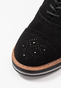 Chaussure oxford en suède noir avec des détails perforés et des accents argentés, dotée d'une semelle intermédiaire en cuir tan et d'une semelle extérieure en caoutchouc noir.