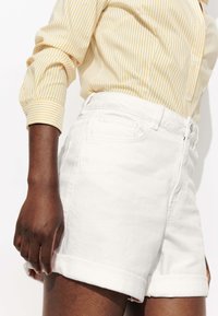 Shorts en denim blanc avec des ourlets retroussés, dotés d'une taille haute, d'une fermeture à bouton à l'avant et de poches à l'arrière. Assortis à une chemise à rayures jaunes.