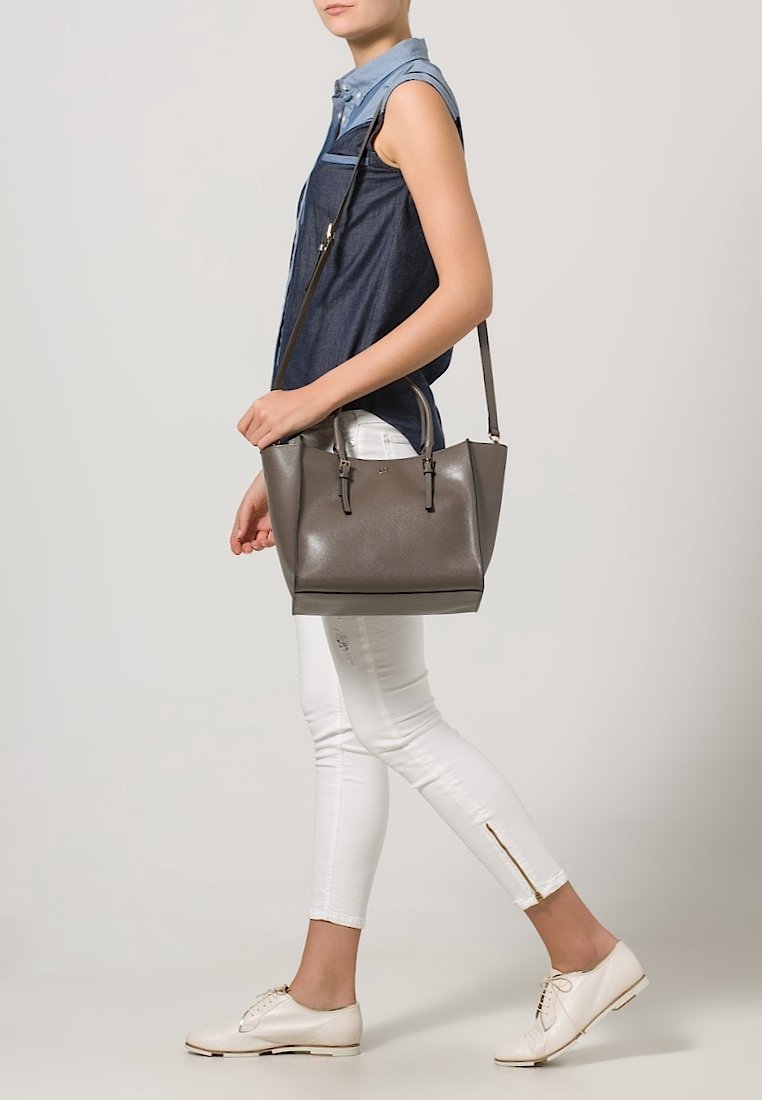 Personne marchant portant un sac à main en cuir gris moyen avec une bandoulière, vêtue d'une chemise en jean sans manches, d'un pantalon blanc et de chaussures blanches.