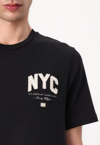 T-shirt en coton noir à manches courtes. Présente un design brodé "NYC" en crème et un texte supplémentaire en dessous, avec un col rond classique.