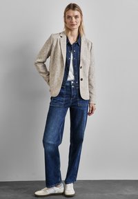 Hellgrauer strukturierter Blazer über einem Jeanshemd, kombiniert mit hochgeschnittenen dunkelblauen Jeans und weißen Sneakern. Sichtbare schlichte Knopfdetails.