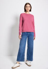 Giovane donna che indossa un maglione rosa, jeans blu a gamba larga e scarpe da tennis bianche, in piedi davanti a uno sfondo grigio chiaro uniforme.