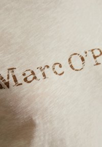 Camiseta de algodón color beige claro con una superficie ligeramente texturada, que presenta el texto impreso "Marc O'Polo" en una fuente marrón y envejecida.